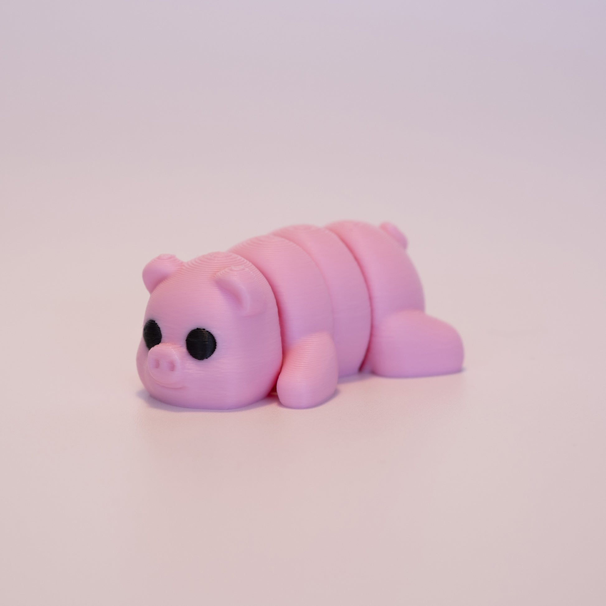 A 3D printed Pig figurine laying on its stomach over an off white background. The figurine is entirely pink with round black eyes.