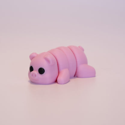 A 3D printed Pig figurine laying on its stomach over an off white background. The figurine is entirely pink with round black eyes.