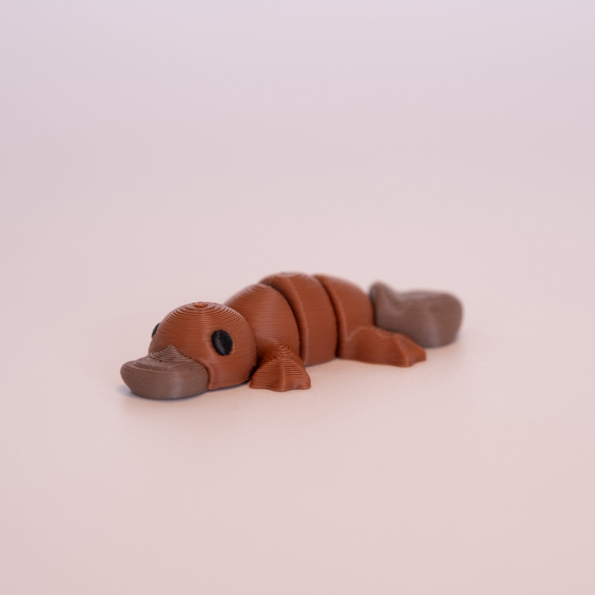 A 3D printed Platypus figurine laying on its stomach over an off white background. The figurine is mostly a warm brown and has round black eyes, with a cool brown beak and tail.