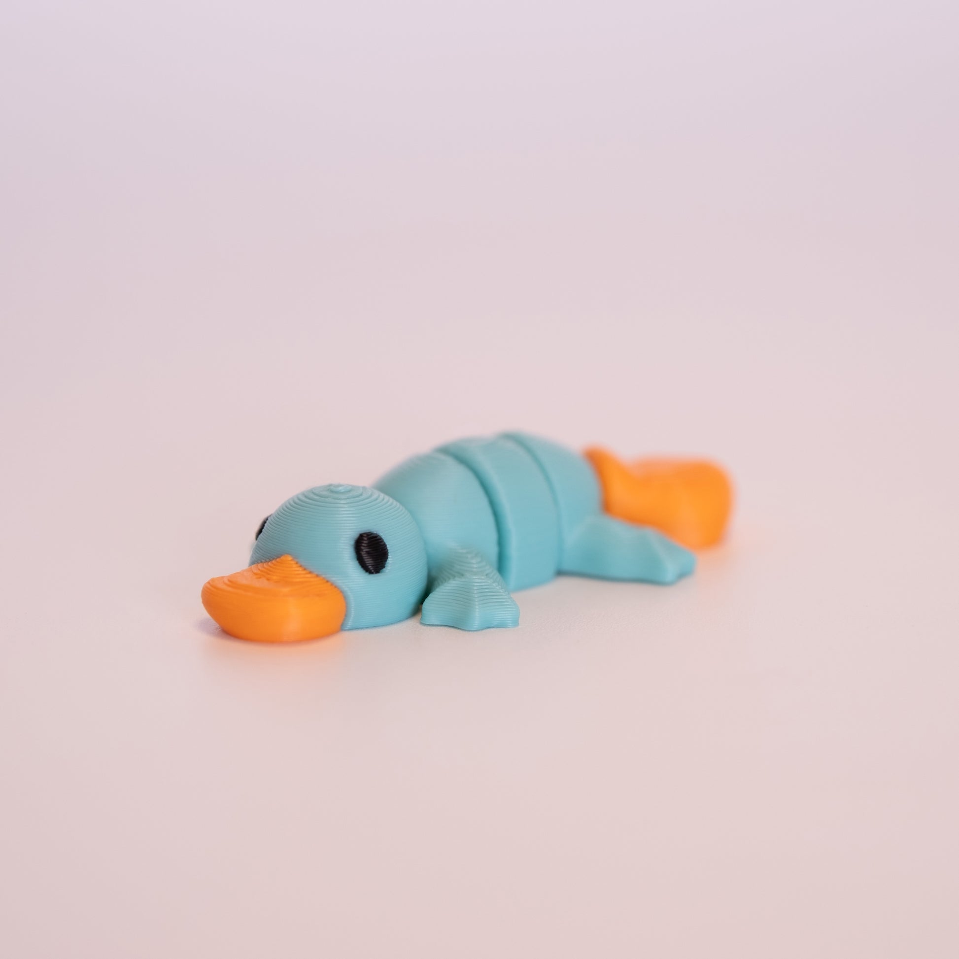 A 3D printed Platypus (Teal) figurine laying on its stomach over an off white background. The figurine is mostly teal and has round black eyes, with an orange beak and tail.