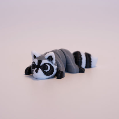 A 3D printed Raccoon figurine laying on its stomach over an off white background. The figurine is mostly grey with black paws, white scleras, and black iris/pupils looking up. The Raccoon also has white and black: ears, face, and striped tail.