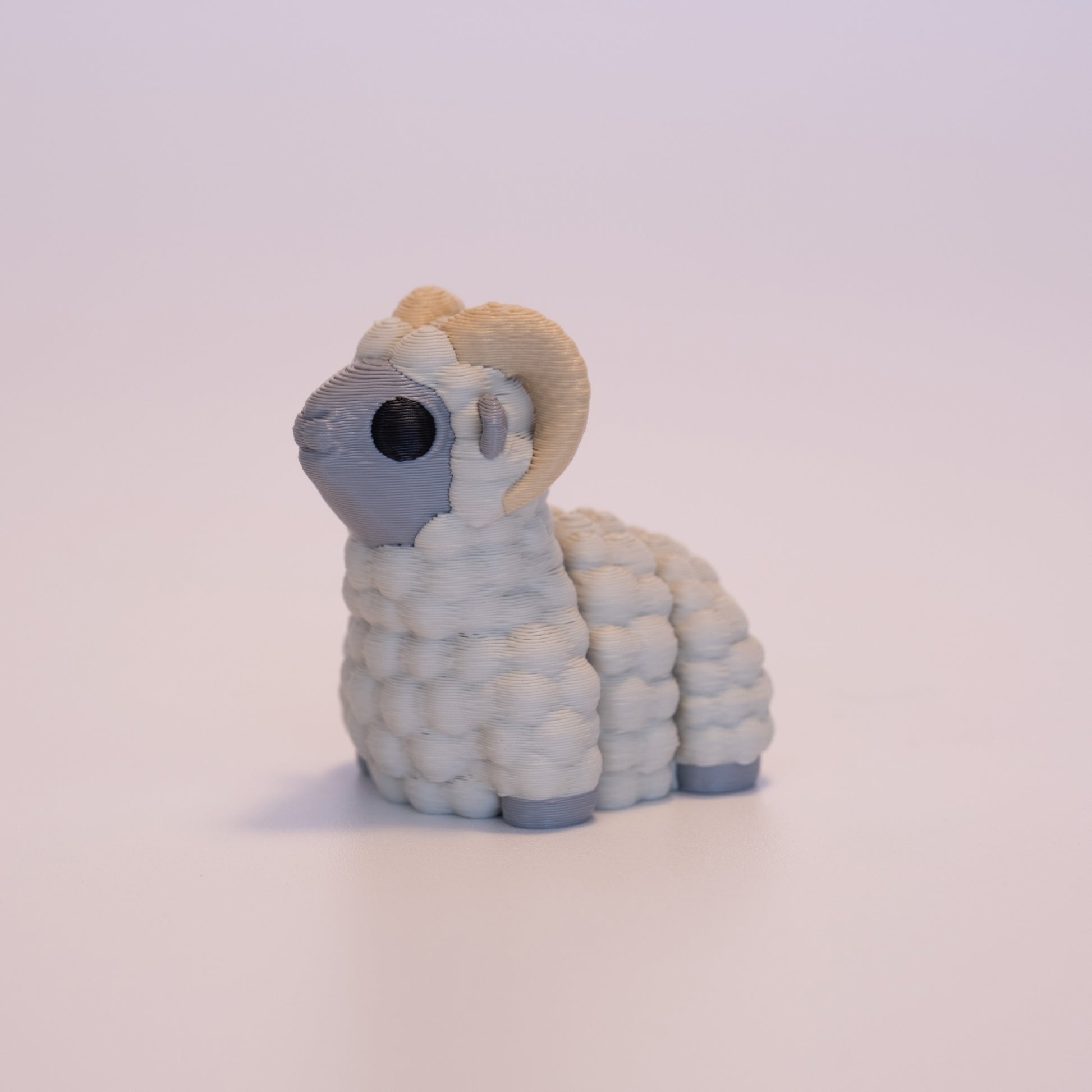 A 3D Printed Ram figurine standing on its 4 legs on an off white background. The Ram’s body is grey with muted beige wool, round black eyes, and tan horns. 