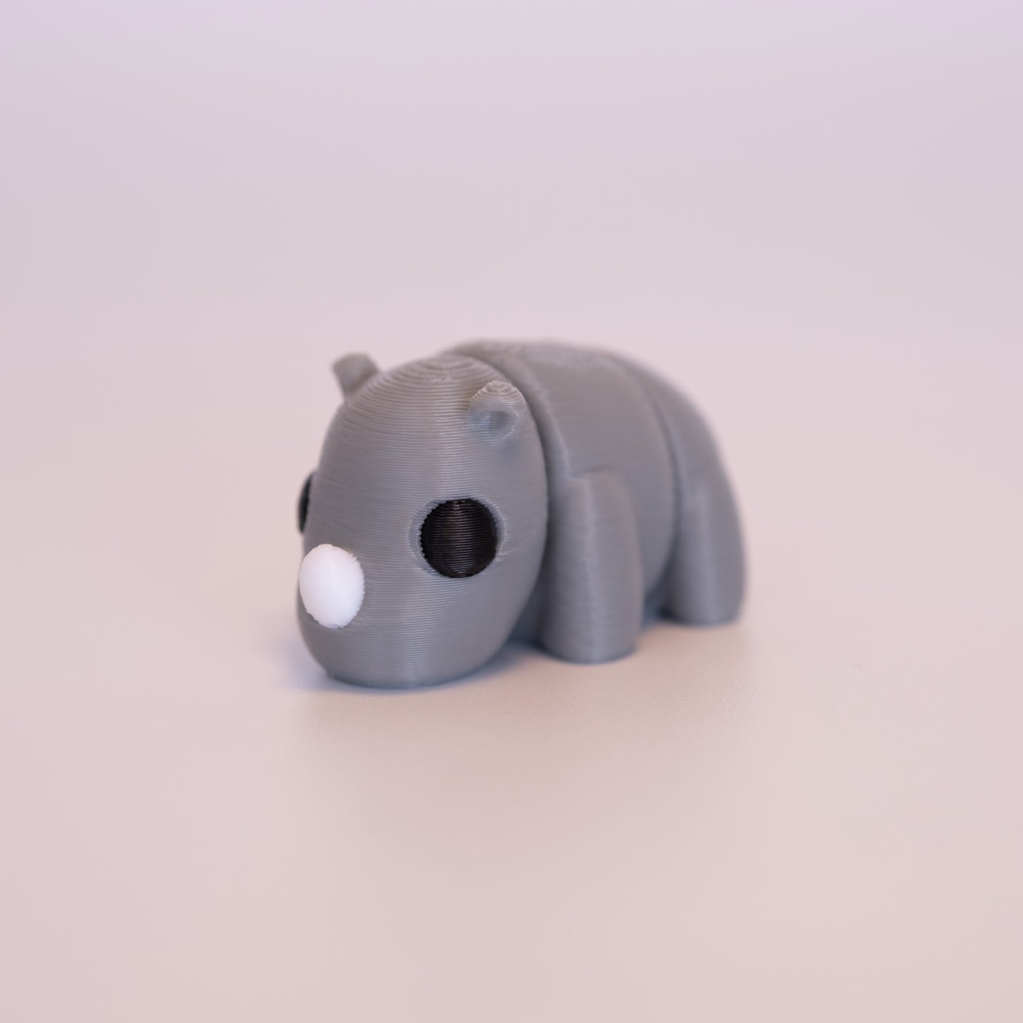 A 3D Printed Rhino figurine standing on its 4 legs on an off white background. The Rhino is mostly grey with round black eyes, and a white horn. 