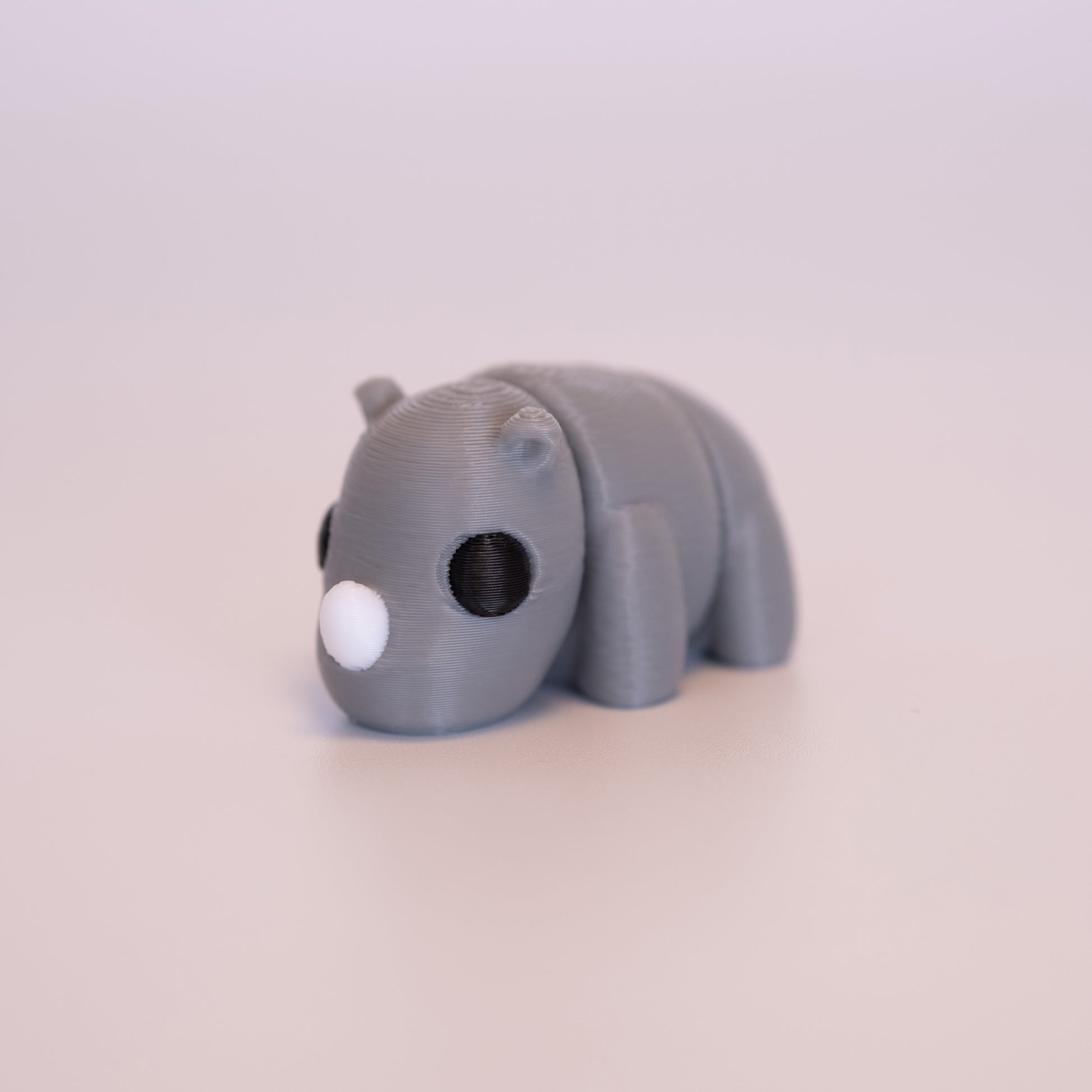 A 3D Printed Rhino figurine standing on its 4 legs on an off white background. The Rhino is mostly grey with round black eyes, and a white horn. 
