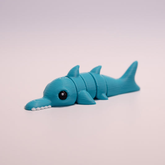 A 3D printed Sawshark figurine laying on its stomach over an off white background. The figurine is mostly teal, round black eyes with a small white accent dot in each eye, and white teeth.