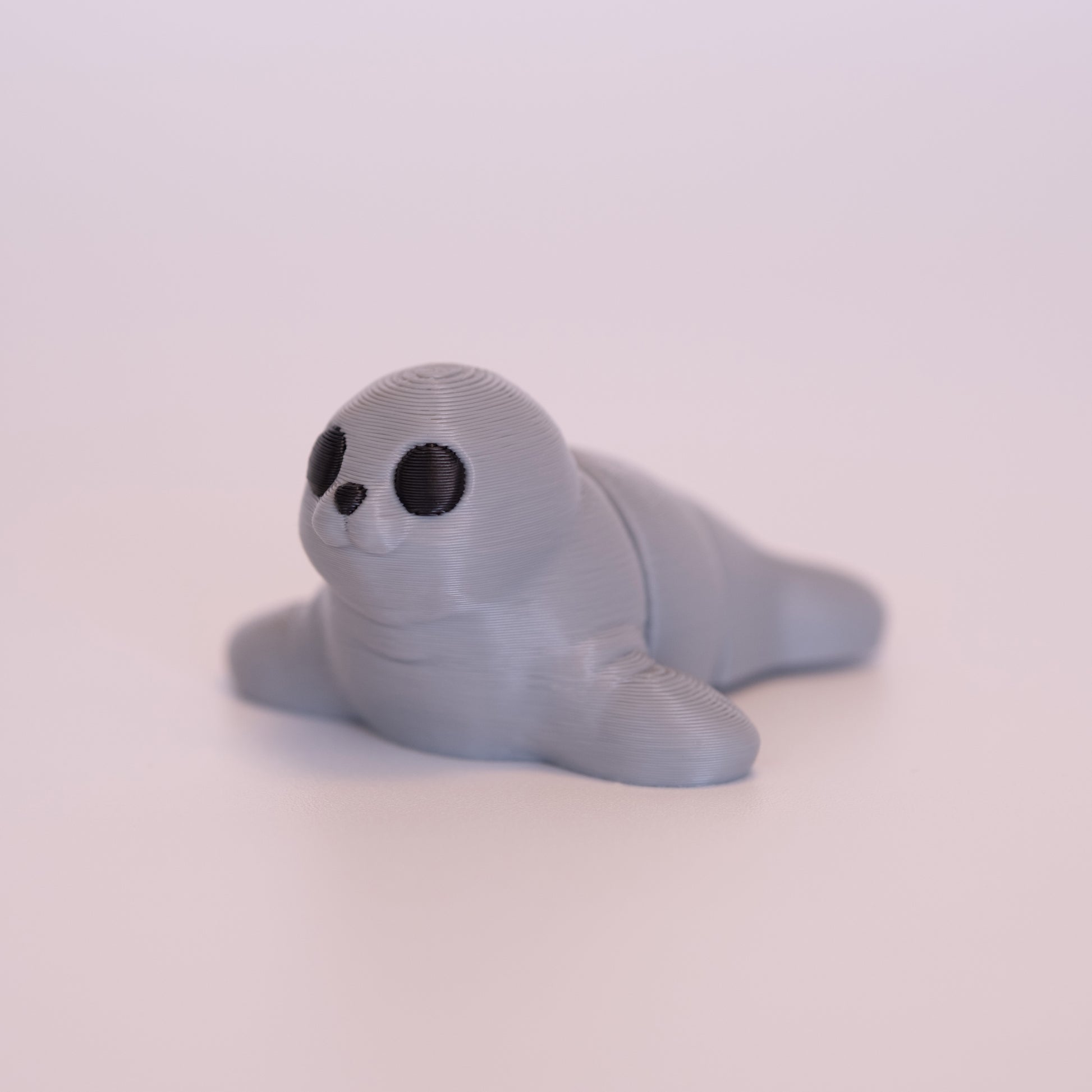 A 3D printed Seal figurine laying on its stomach over an off white background. The figurine is entirely light grey with round black eyes and a black nose.
