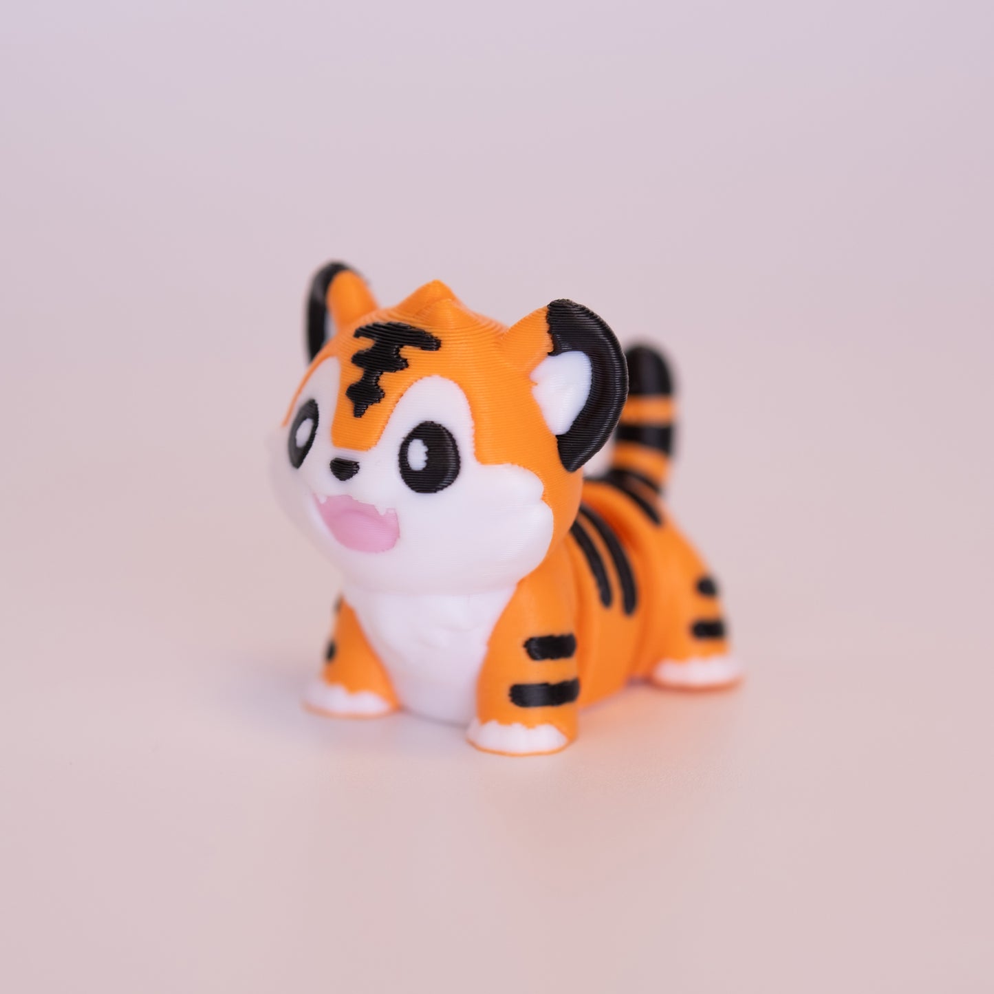 A 3D Printed Tiger figurine standing on its 4 legs on an off white background. The Tiger is mostly orange with white and black accents throughout. It has round black eyes, each with 1 white accent dot in each eye, a black nose, and pink mouth. 
