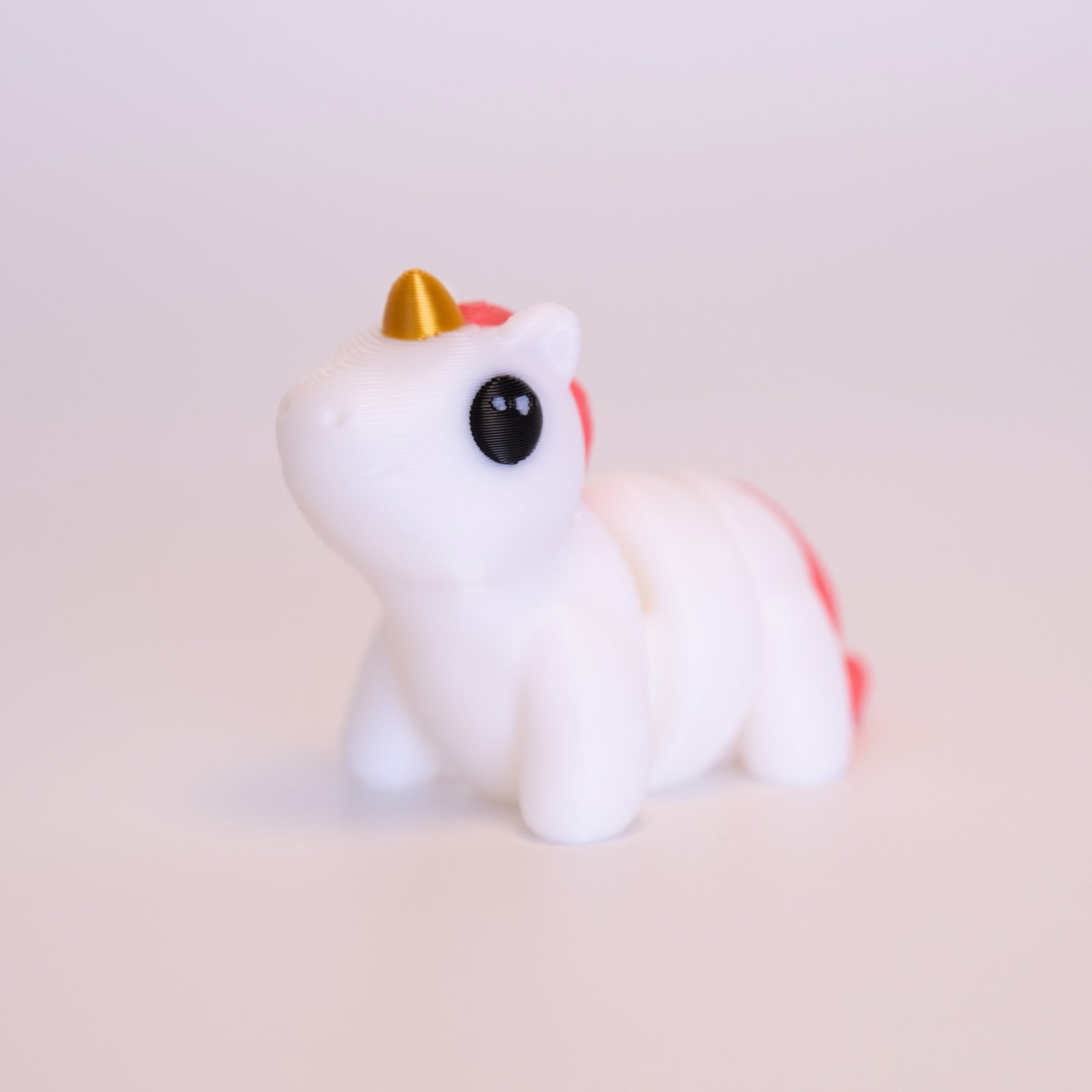 A 3D Printed Unicorn figurine standing on its 4 legs on an off white background. The Unicorn is mostly white, with a gold horn, pink mane and tail, and round black eyes with 2 small white accent dots in each eye.