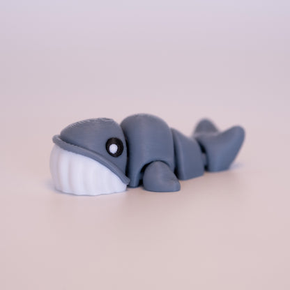 A 3D printed Blue Whale figurine laying on its stomach over an off white background. The figurine is mostly blue-grey, round black eyes with a white dot accent in each eye, and white bottom half of the face.
