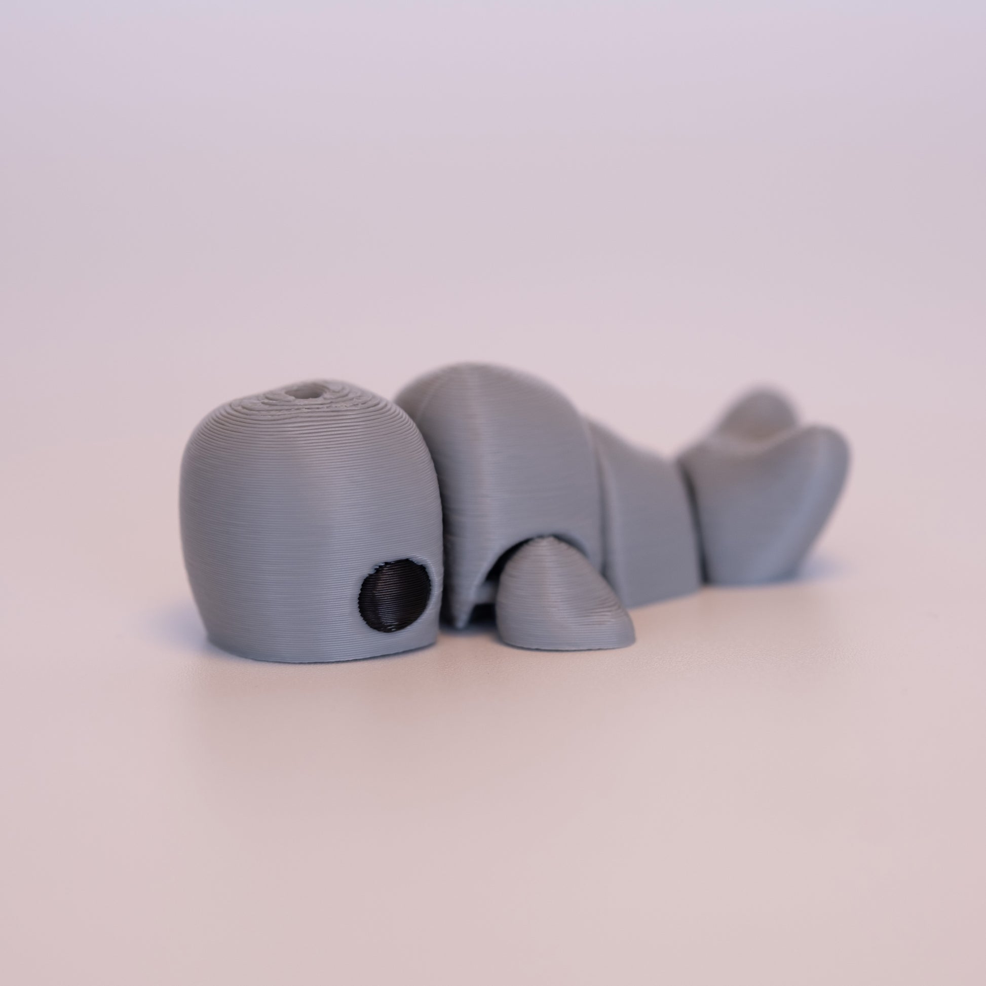 A 3D printed Sperm Whale figurine laying on its stomach over an off white background. The figurine is entirely light grey with round black eyes.