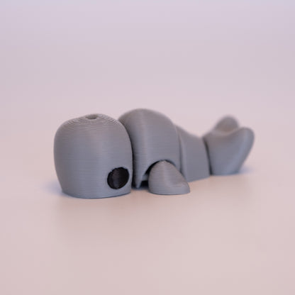 A 3D printed Sperm Whale figurine laying on its stomach over an off white background. The figurine is entirely light grey with round black eyes.