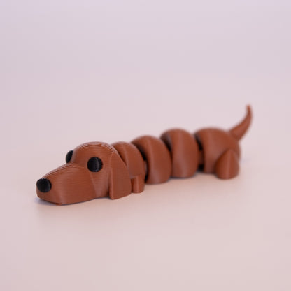 A 3D printed Wiener Dog figurine laying on its stomach over an off white background. The figurine is entirely a warm brown with round black eyes and a black nose.